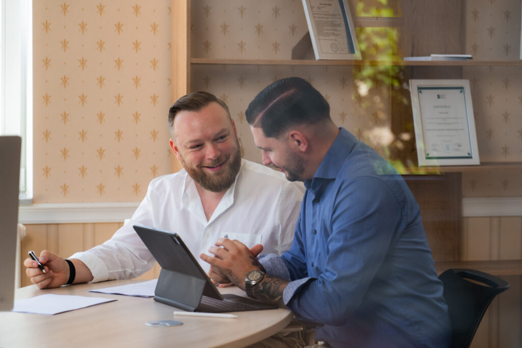 Zwei Männer sitzen lachend an einem Tisch und arbeiten gemeinsam an einem Tablet.
