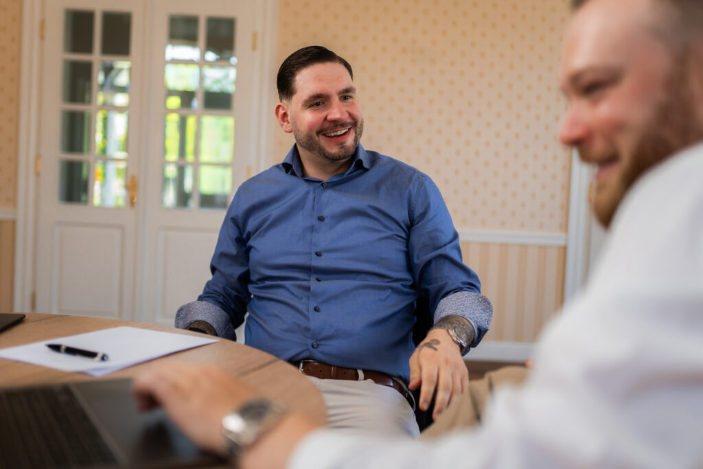 Das Bild zeigt zwei Männer in einem hell eingerichteten Raum bei einem entspannten Gespräch, wobei der Mann im blauen Hemd lächelnd im Mittelpunkt sitzt.