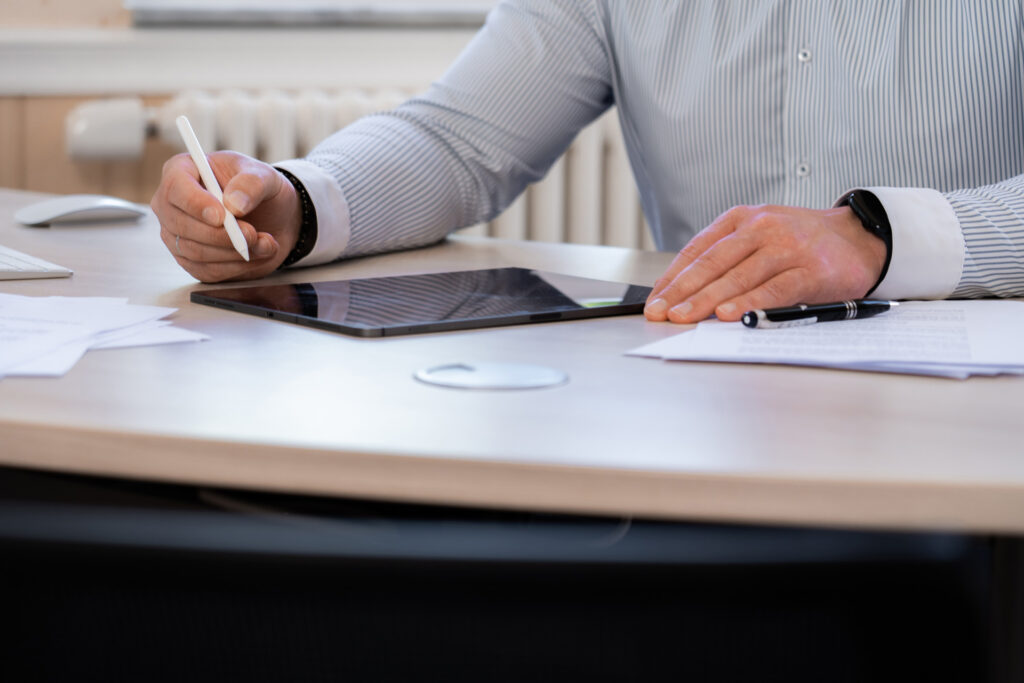 Ein Mann arbeitet mit einem Tablet und Stift an einem Schreibtisch, auf dem auch Dokumente und ein Stift liegen.